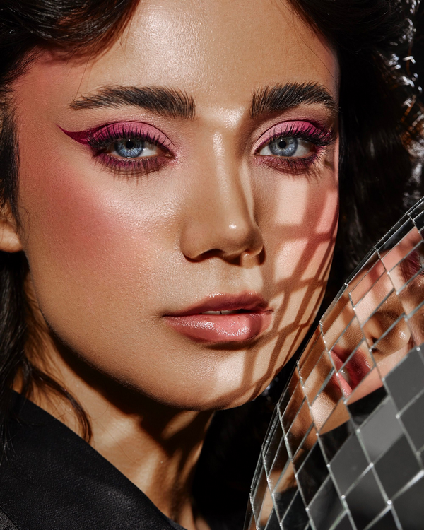 Asian model in an editorial beauty shot with bold magenta-pink winged liner, sculpted cheekbone and a small mirror grid catching the light — a finished dimensional glam look on Asian features